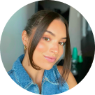 Close-up portrait of a young woman with dark hair tied back, wearing a blue denim jacket and gold hoop earrings, smiling softly.