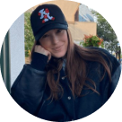 Smiling young woman with long hair wearing a black jacket and a black baseball cap with a red and white figure on it, resting her head on her right hand outdoors near a potted plant.