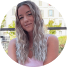 Smiling young woman with long wavy blonde hair wearing a pink top, outdoors with buildings and greenery.