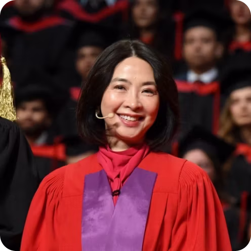 Smiling woman with black hair wearing a red academic gown and a purple stole with a microphone headset, standing in front of a blurred audience in similar academic attire.