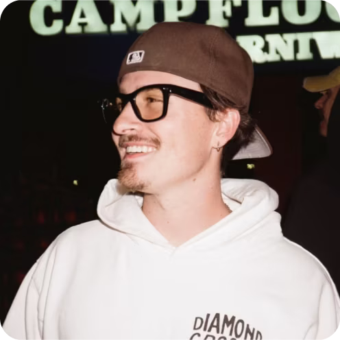 Smiling man wearing a brown baseball cap backwards, black glasses, and a white hoodie with text on it, standing in front of partially visible illuminated signage.
