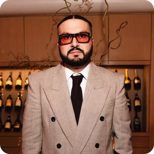 Man with a beard and mustache wearing large amber-tinted sunglasses, a beige double-breasted suit, white shirt, and black tie standing indoors in front of a wooden cabinet with bottles.