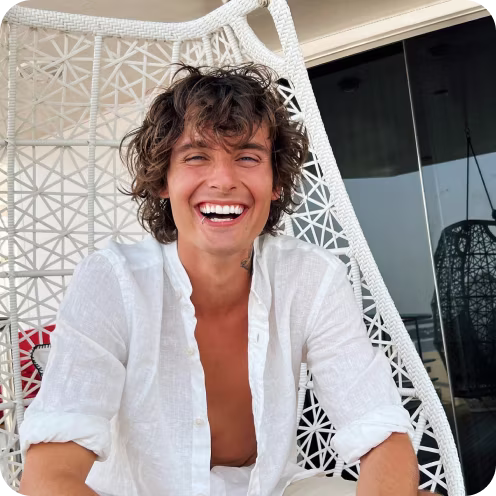 Smiling young man with curly hair wearing an open white shirt sitting in a white woven hanging chair.