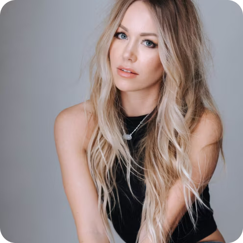 Blonde woman with long wavy hair and blue eyes wearing a black sleeveless top and a silver necklace, looking at the camera.