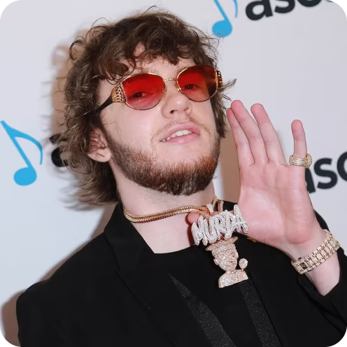 Young man with curly brown hair and beard wearing red-tinted sunglasses and dressed in black, holding up a large diamond-studded necklace pendant that spells 'MORAD' with a figure below, posed in front of a white backdrop with partial logos containing musical notes.
