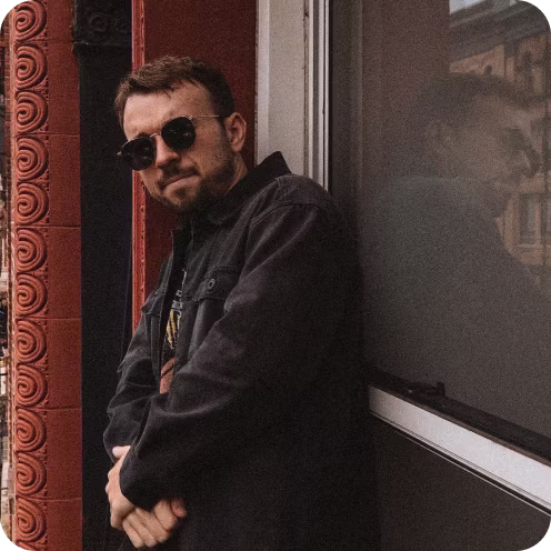 Man wearing round black sunglasses and a dark jacket leaning against a window, with his reflection visible on the glass and an ornate red brick wall behind him.