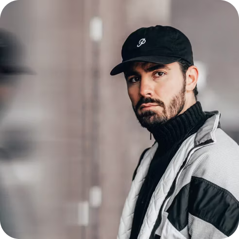 Bearded man wearing a black cap and black and white jacket, looking at the camera with a serious expression.