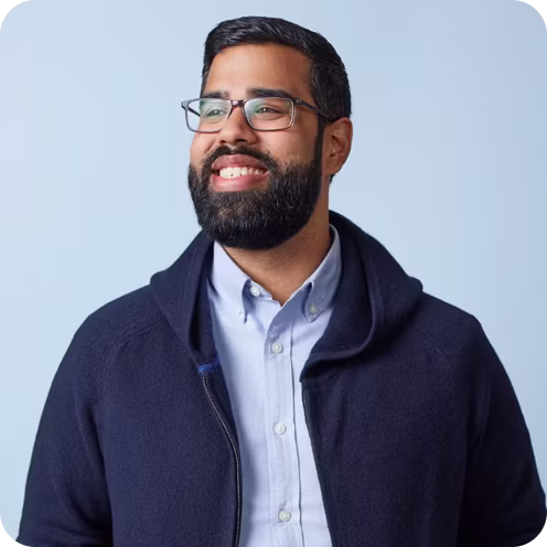 Smiling man with black hair, beard, and glasses wearing a light blue button-up shirt and dark blue jacket.