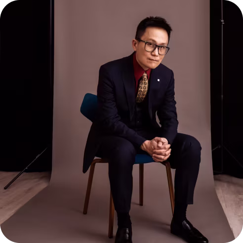 Man wearing black suit, red shirt, and patterned tie sitting on a blue chair against a brown backdrop with hands clasped and looking forward.