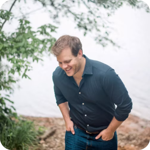 A smiling man with brown hair wearing a dark blue button-up shirt and jeans, leaning slightly forward outdoors near a body of water with greenery.