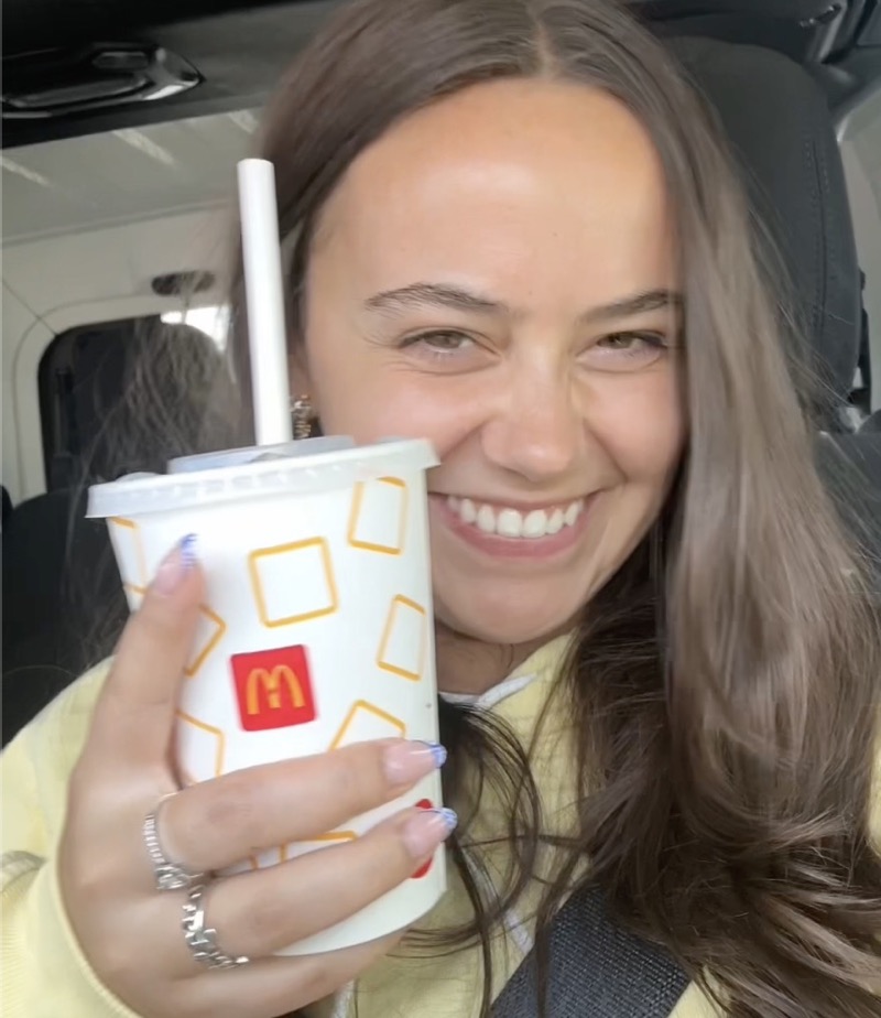 A smiling woman with long brown hair inside a vehicle holding up a McDonald's drink cup with a straw, wearing a yellow top and a seatbelt.