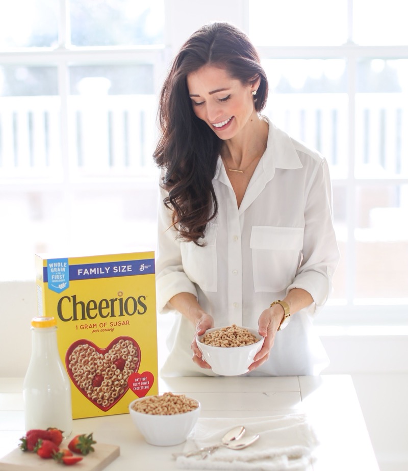 Smiling woman with long brown hair wearing a white shirt holding a bowl of Cheerios cereal in a bright kitchen, with a family-size box of Cheerios, a bottle of milk, strawberries, and another bowl of cereal on the table.