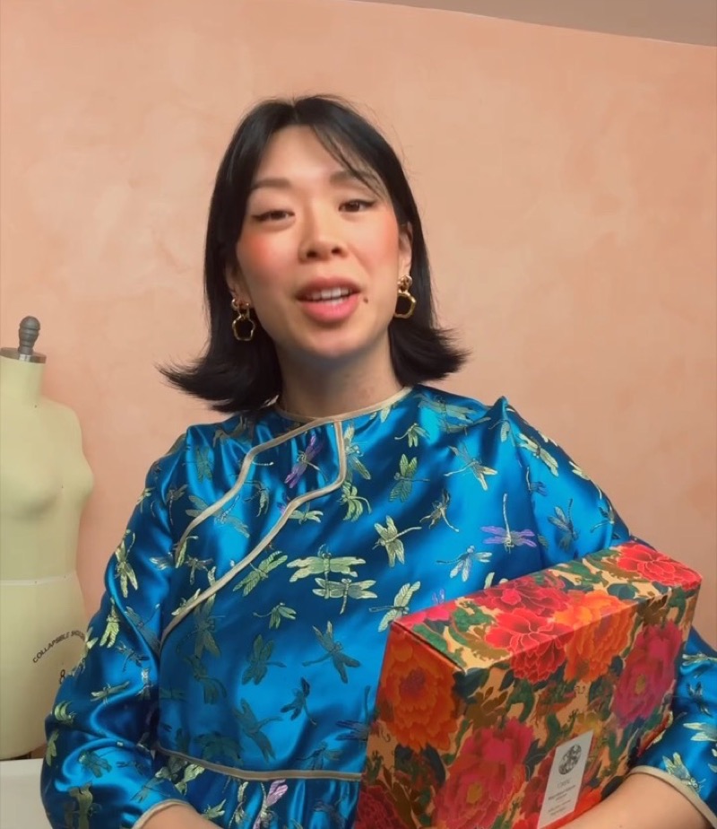 Woman with short black hair wearing a blue satin floral-patterned blouse, holding a colorful rectangular box with a floral design against a beige background and a dress form to her side.