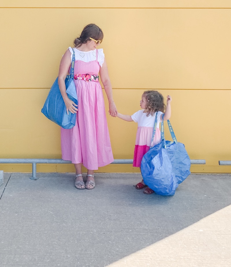 A woman in a pink dress holding hands with a young girl in a pink and white dress against a yellow wall, both carrying large blue reusable shopping bags.