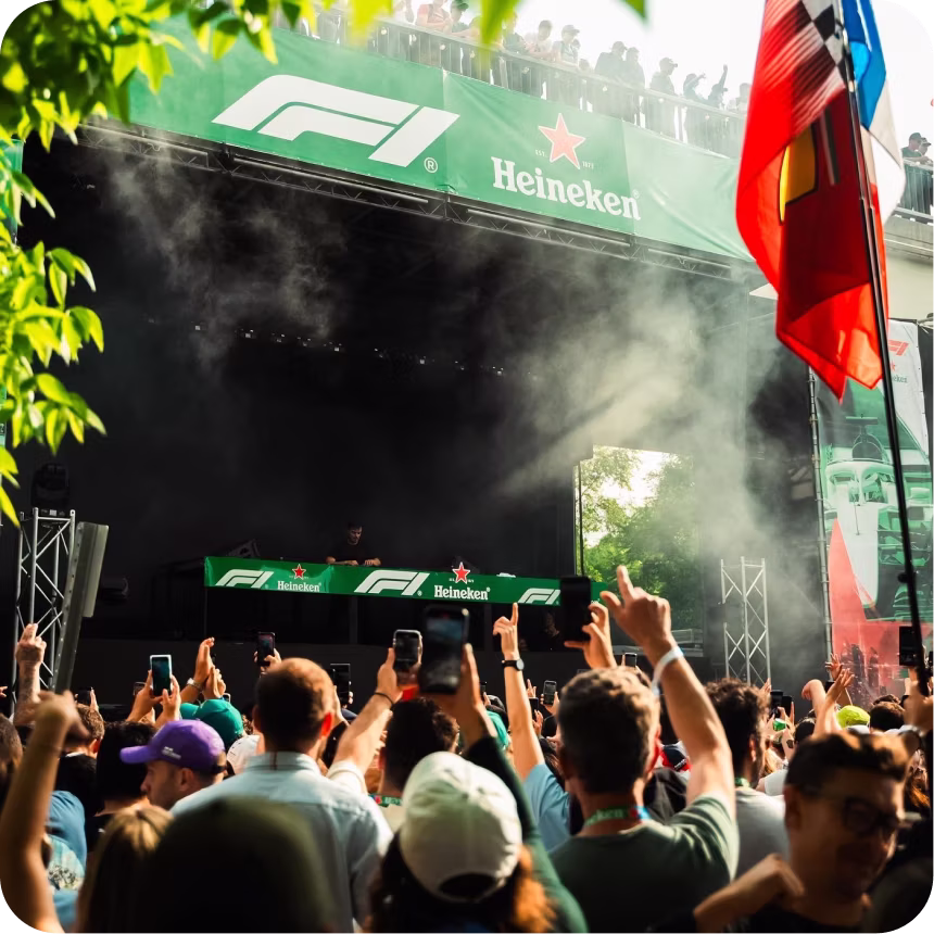 Crowd at an outdoor event raising hands and holding phones in front of a stage with green Heineken and F1 banners, a DJ performing, and a flag waving on the right side.