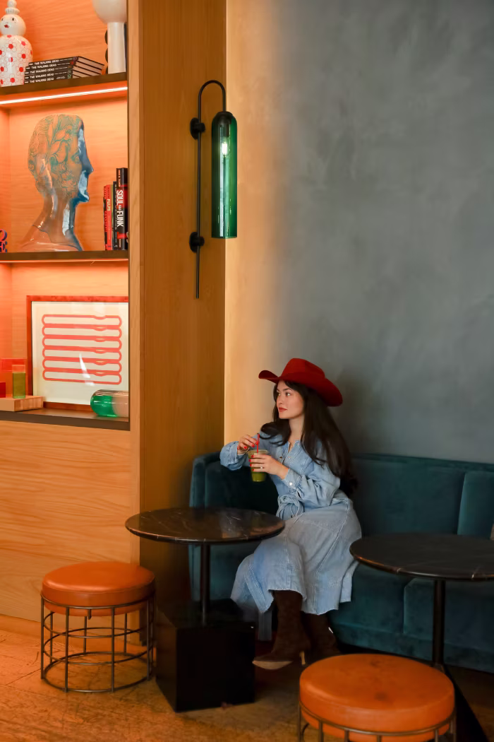 Woman wearing a red hat and blue dress sitting on a dark green velvet couch in a modern cafe corner, holding a drink with a straw, next to two small round tables and two orange stools, with wooden shelves and green wall light in the background.