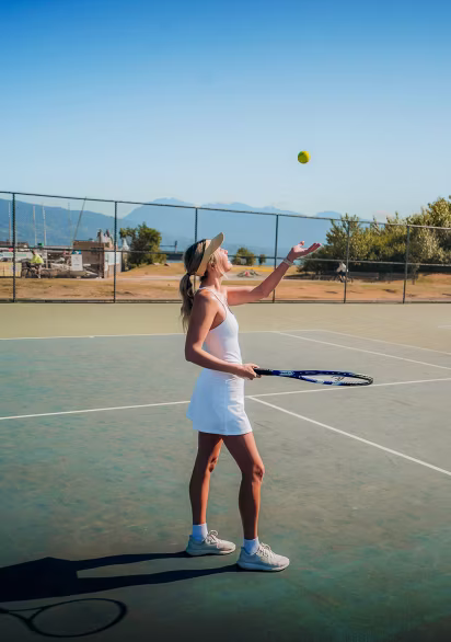 Woman in a white tennis dress and visor tossing a tennis ball up with one hand and holding a tennis racket with the other on a sunny outdoor tennis court.