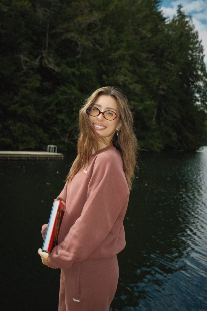 Smiling young woman with glasses wearing a matching mauve sweatshirt and pants, holding books, standing by a lake surrounded by dense green forest under a partly cloudy sky.