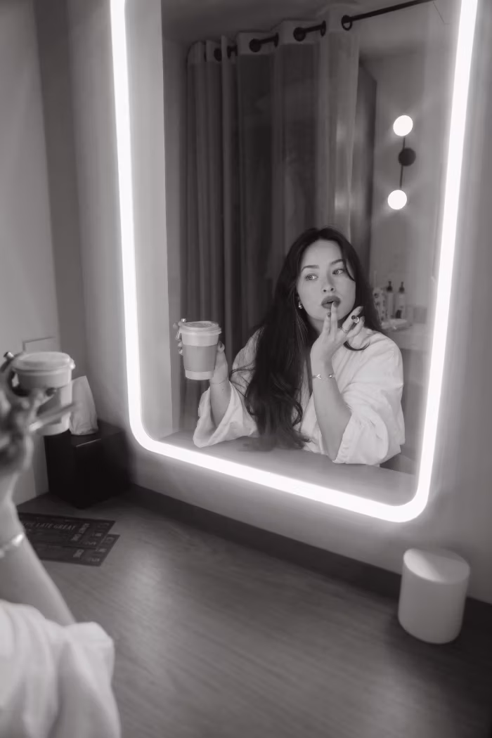 Black and white photo of a woman in a bathrobe holding a coffee cup and applying lip balm while looking into a rectangular illuminated mirror in a modern bathroom.
