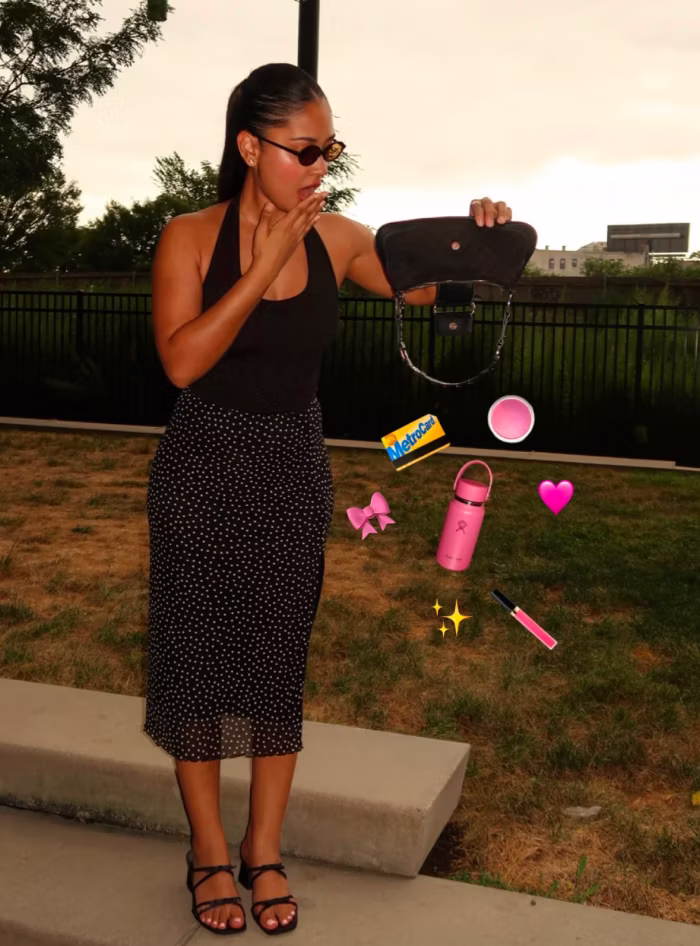 Woman wearing sunglasses, a black sleeveless top, and a black skirt with white polka dots stands on a sidewalk near grass. She holds a small black purse open, as various pink-themed items like a water bottle, lip gloss, bow, and heart float in the air beside her, along with a MetroCard and sparkle emojis.