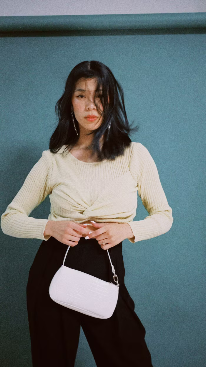 Woman with shoulder-length black hair wearing a light yellow ribbed sweater and black pants, holding a small white handbag against a teal background.