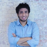 Smiling young man with dark curly hair and beard wearing a light blue button-up shirt, standing with arms crossed against a light brick wall.