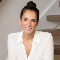 Smiling woman with dark hair tied in a bun, wearing a white shirt and sitting indoors near wooden stairs.