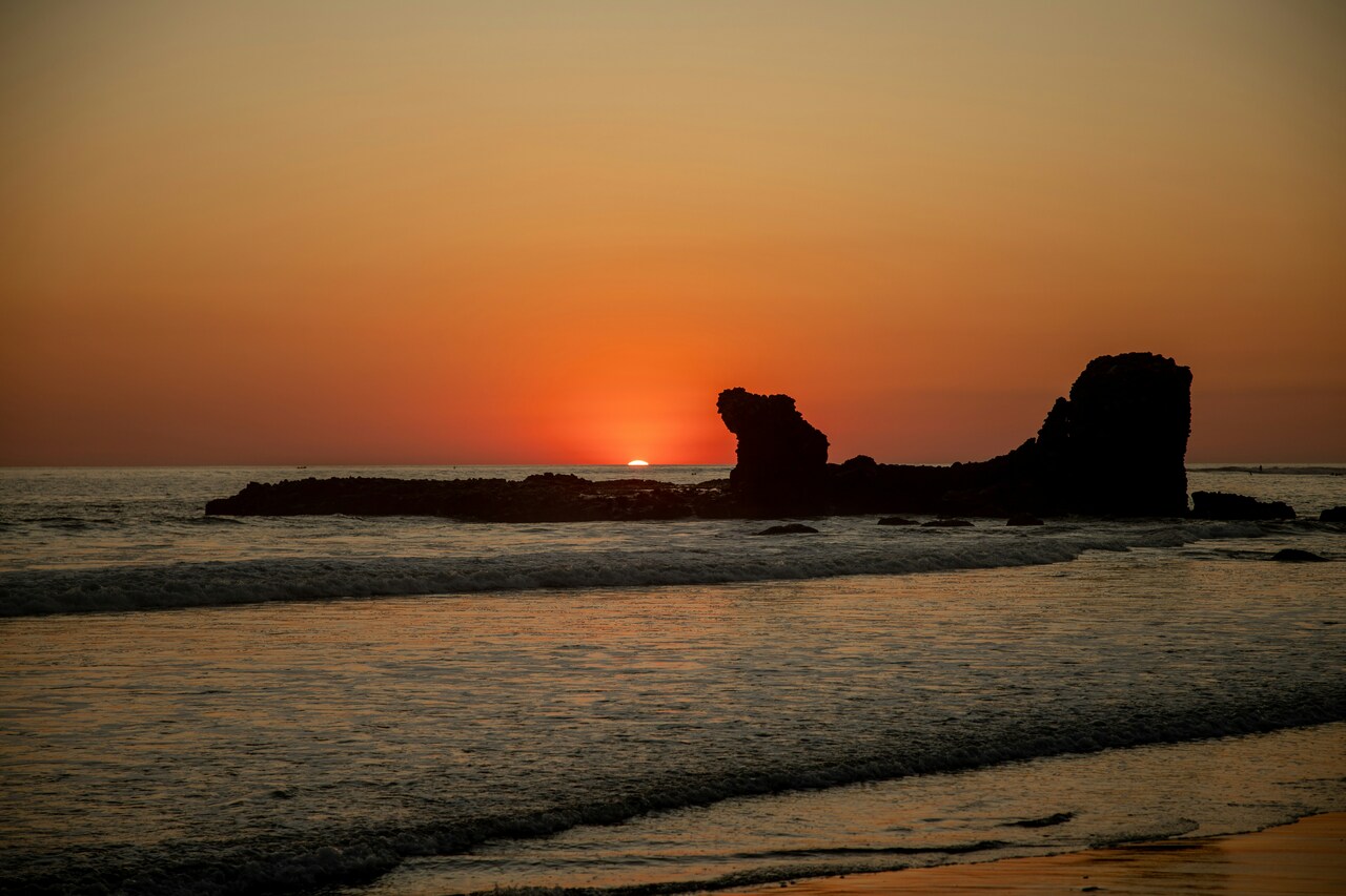 Vivid sunset over the ocean with waves gently washing onto the shore and a large rock formation silhouetted against the orange sky.
