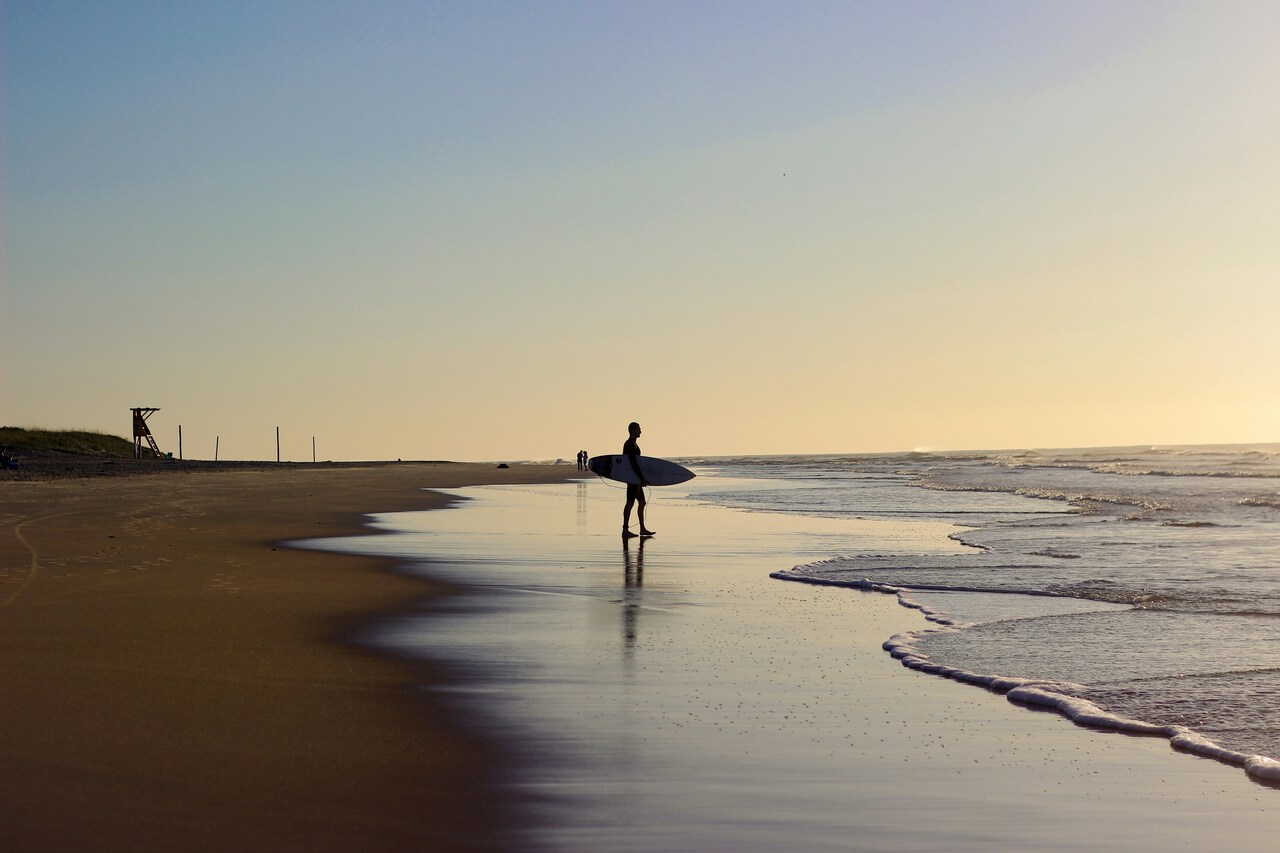 Surfer holding a surfboard at sunset on a quiet beach, with gentle waves washing up on the shore.