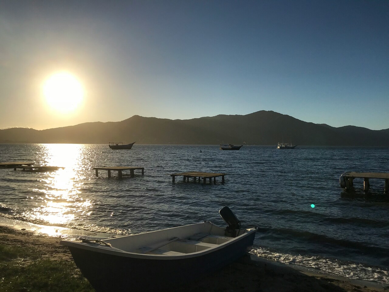 Small boat on a calm shoreline at sunset with wooden piers and distant sailboats on the water surrounded by mountains.
