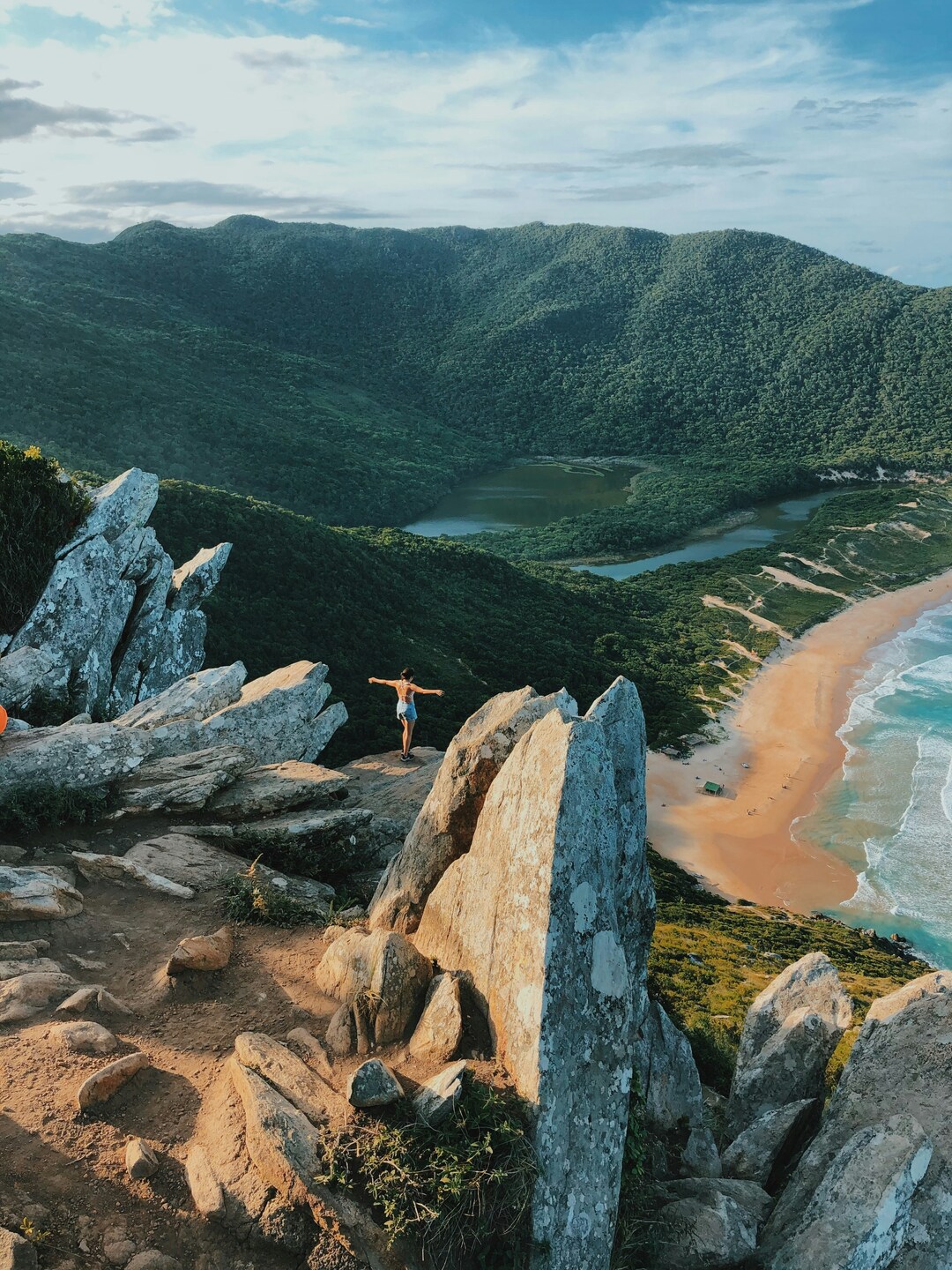 Person standing with arms outstretched on rocky cliffs overlooking a scenic beach, forested mountains, and a lagoon below.