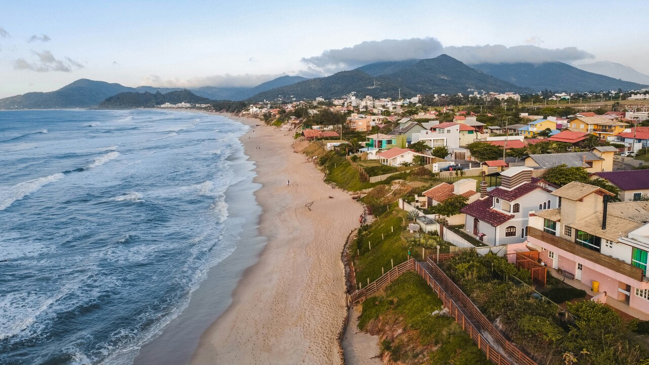 Aerial view of a coastal town with colorful houses along a long sandy beach bordered by ocean waves and green mountains in the background.