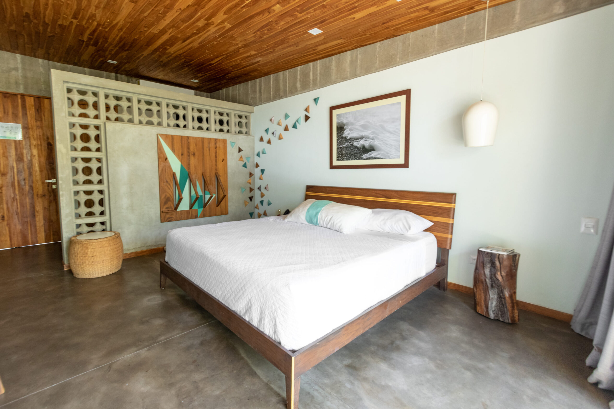 Spacious bedroom with a wooden bed frame, white bedding, artistic wall design, and rustic decor featuring wood accents and natural textures.