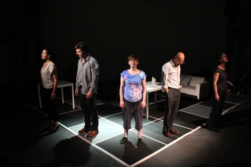 Five performers stand facing forward on the corner of a giant black and white chess board. The background is dark but white tables and a white sofa are positioned on the chess board