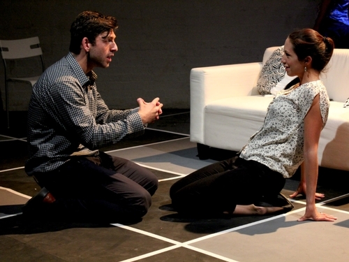 A young man and woman kneel on a floor covered in a large grid of black and white squares, in front of a white sofa. His hands are clasped and she is leaning back
