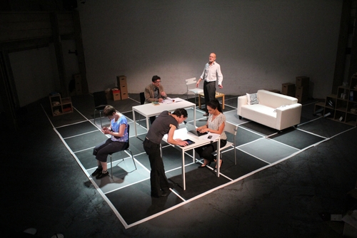There is a huge huge black and white chessboard with tables, chairs and a sofa positioned on different squares. Five characters sit or stand at different angles engrossed in their work