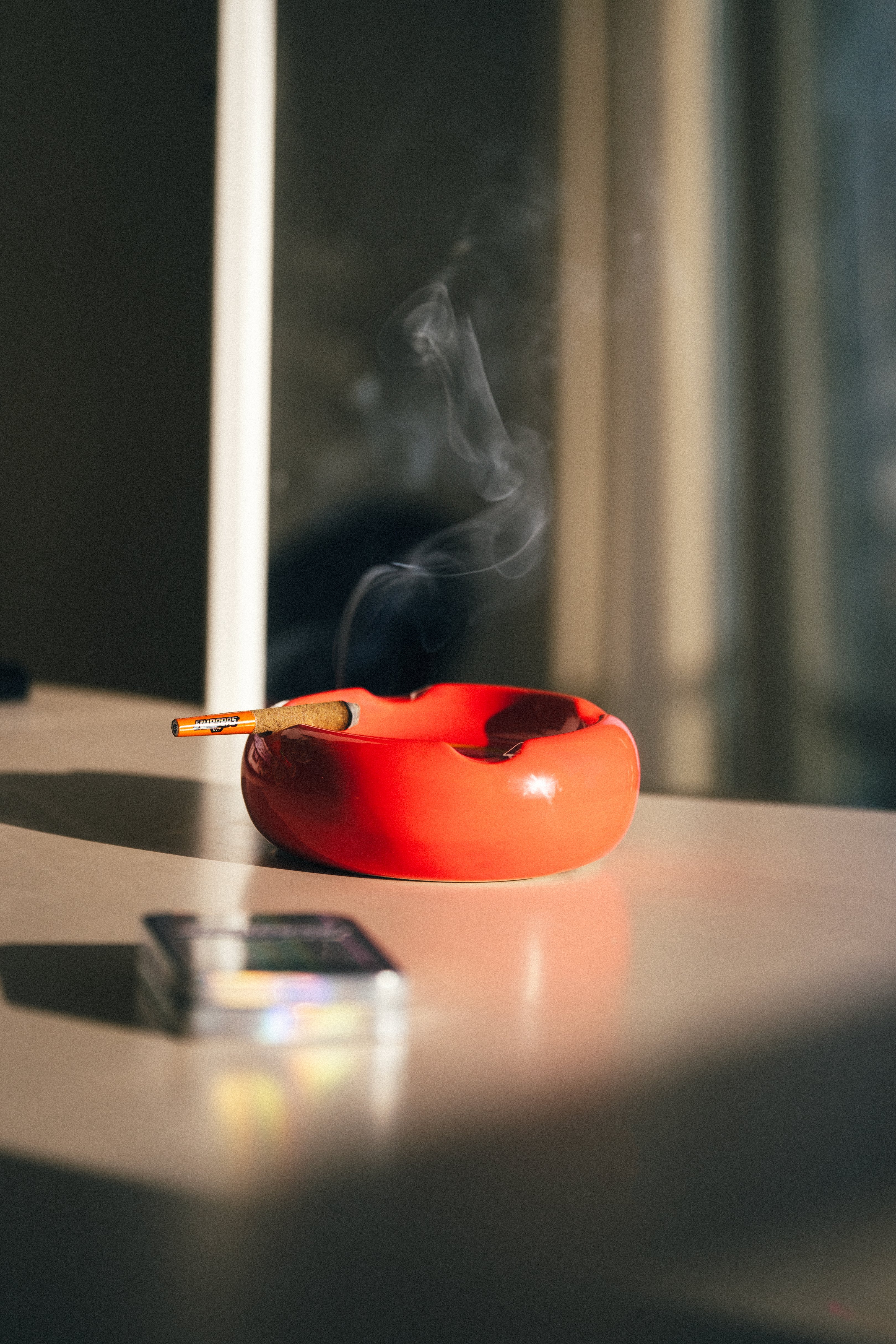 A lit Sluggers pre-roll sitting in an orange round ashtray billowing smoke