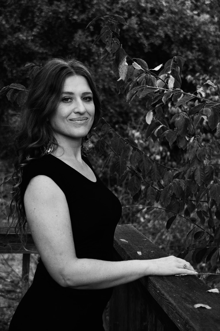 Black-and-white portrait of Maya Elisabeth with long wavy hair standing outdoors beside leafy branches, wearing a sleeveless dark top and looking at the camera.
