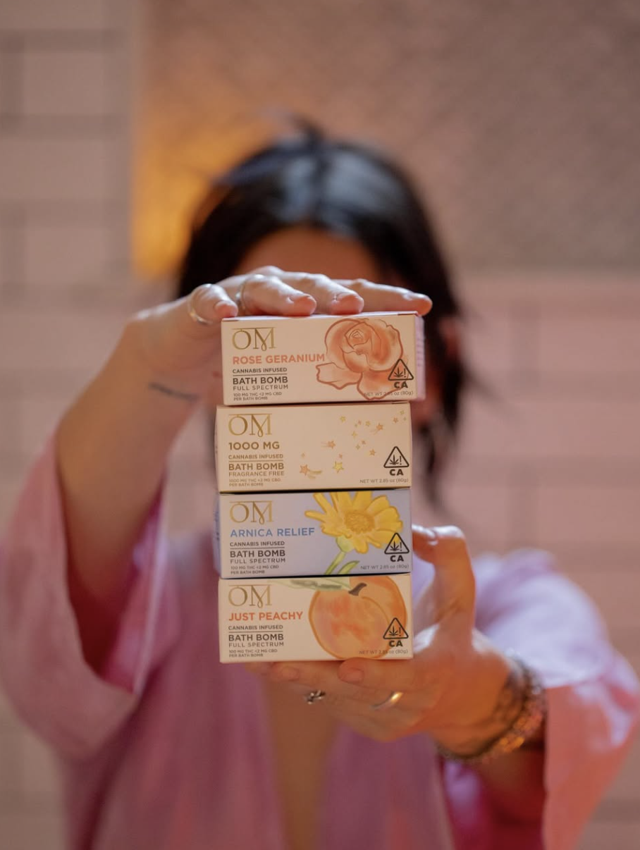 A person in a pink robe holds a stack of four boxed cannabis-infused bath bombs labeled OM Rose Geranium, 1000 MG, Arnica Relief, and Just Peachy, each with floral or fruit illustrations and a California cannabis symbol.