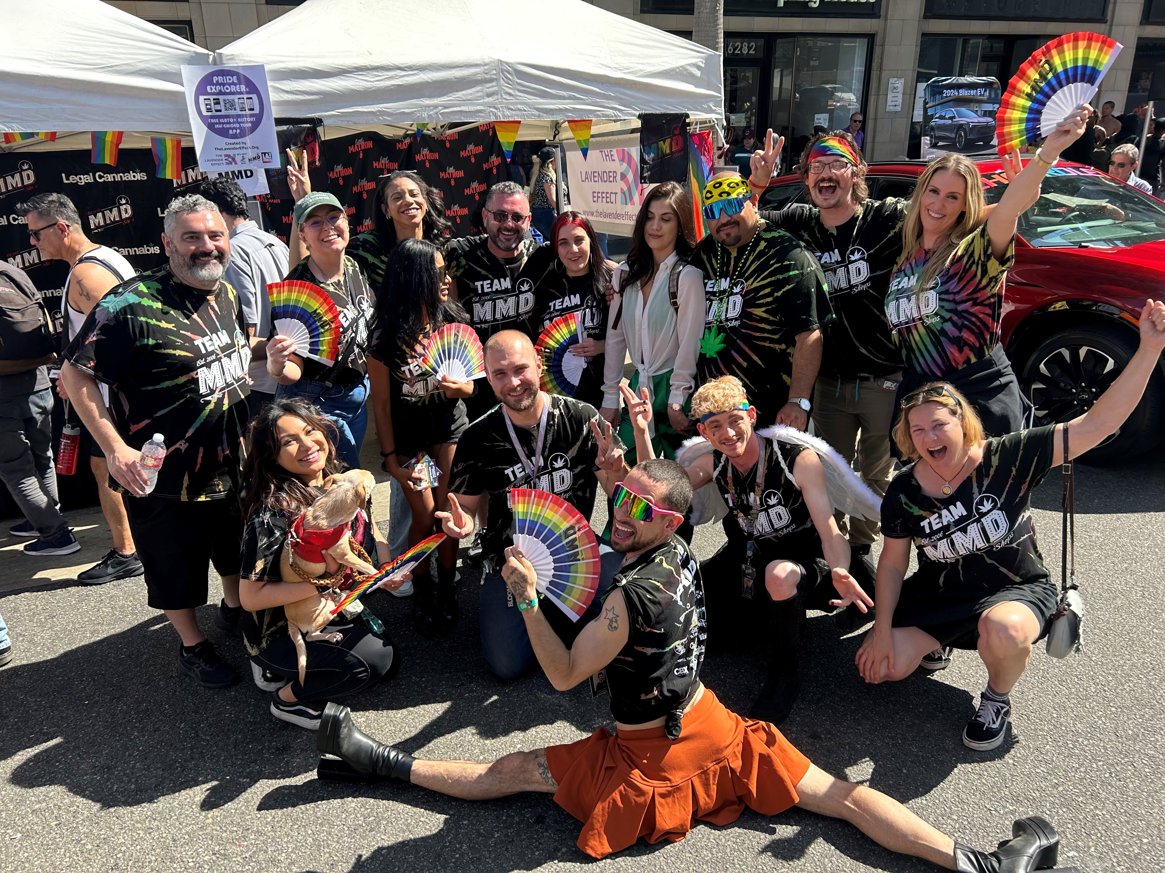 MMD employees in tie-dye "Team MMD" shirts wave rainbow fans at the company's booth during a Pride street festival.
