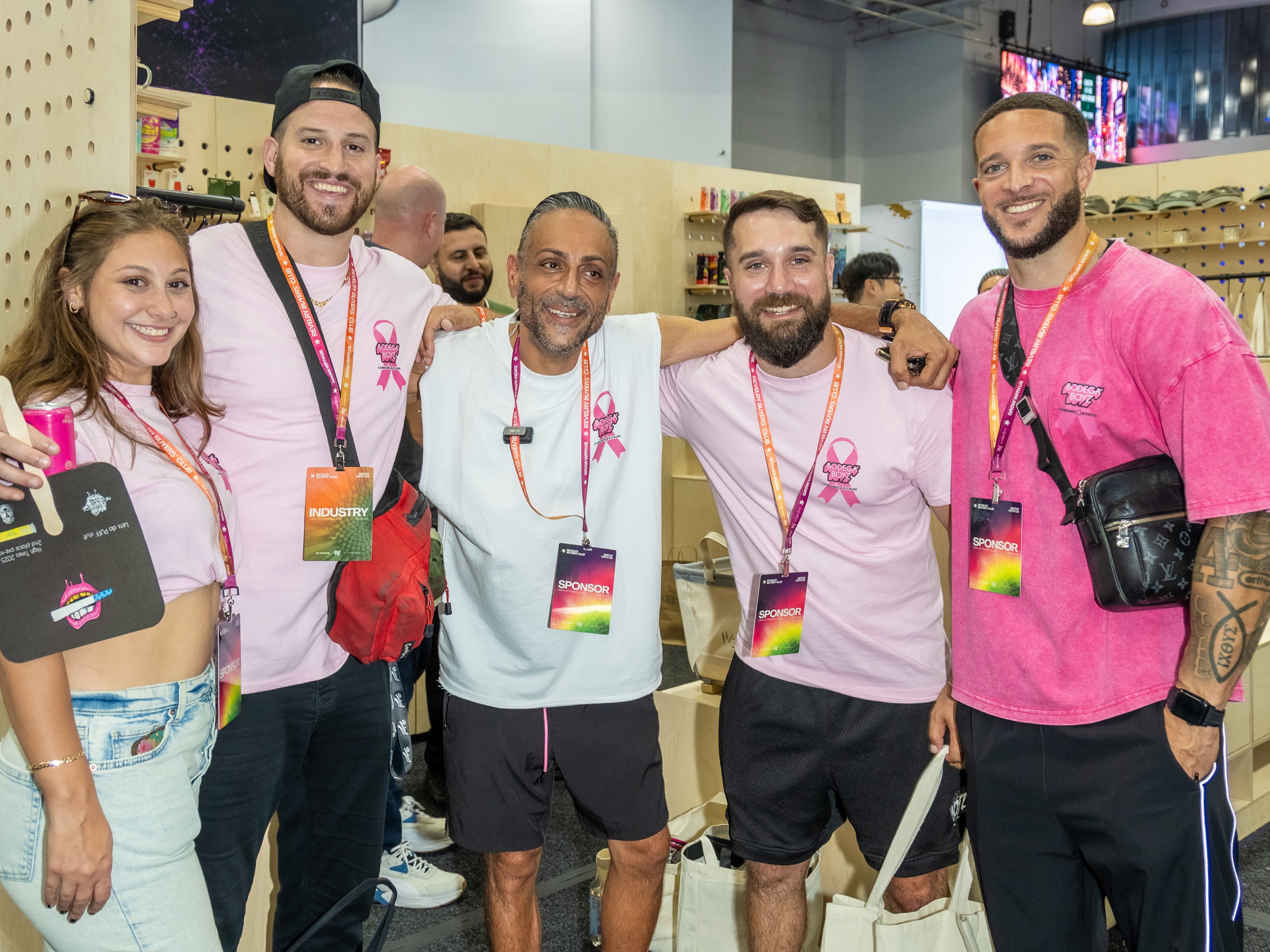 Five members of the Bodega Boyz cannabis team in pink Cannabis 4 A Cause ribbon shirts with sponsor lanyards at a Revelry NYC 2026 booth.