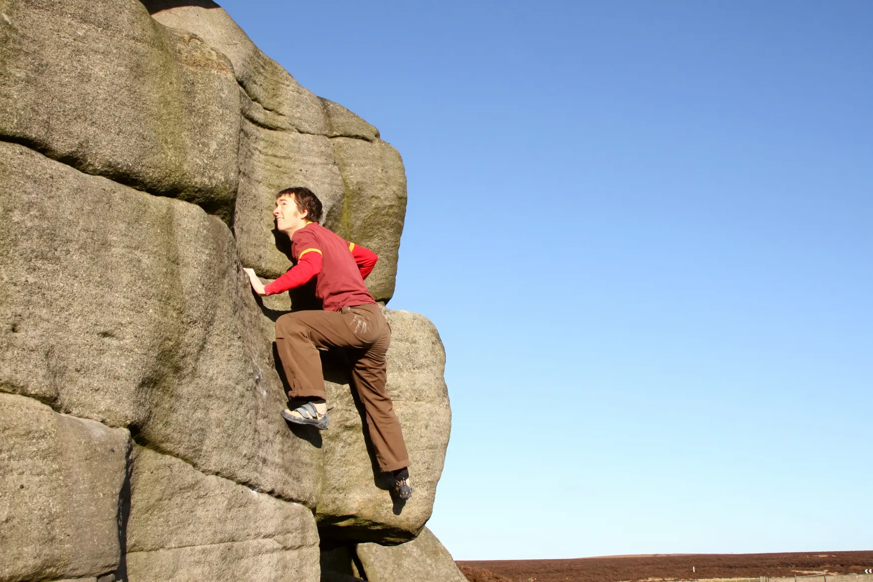 Person wearing a red shirt and brown pants bouldering on a large rock under a clear blue sky.