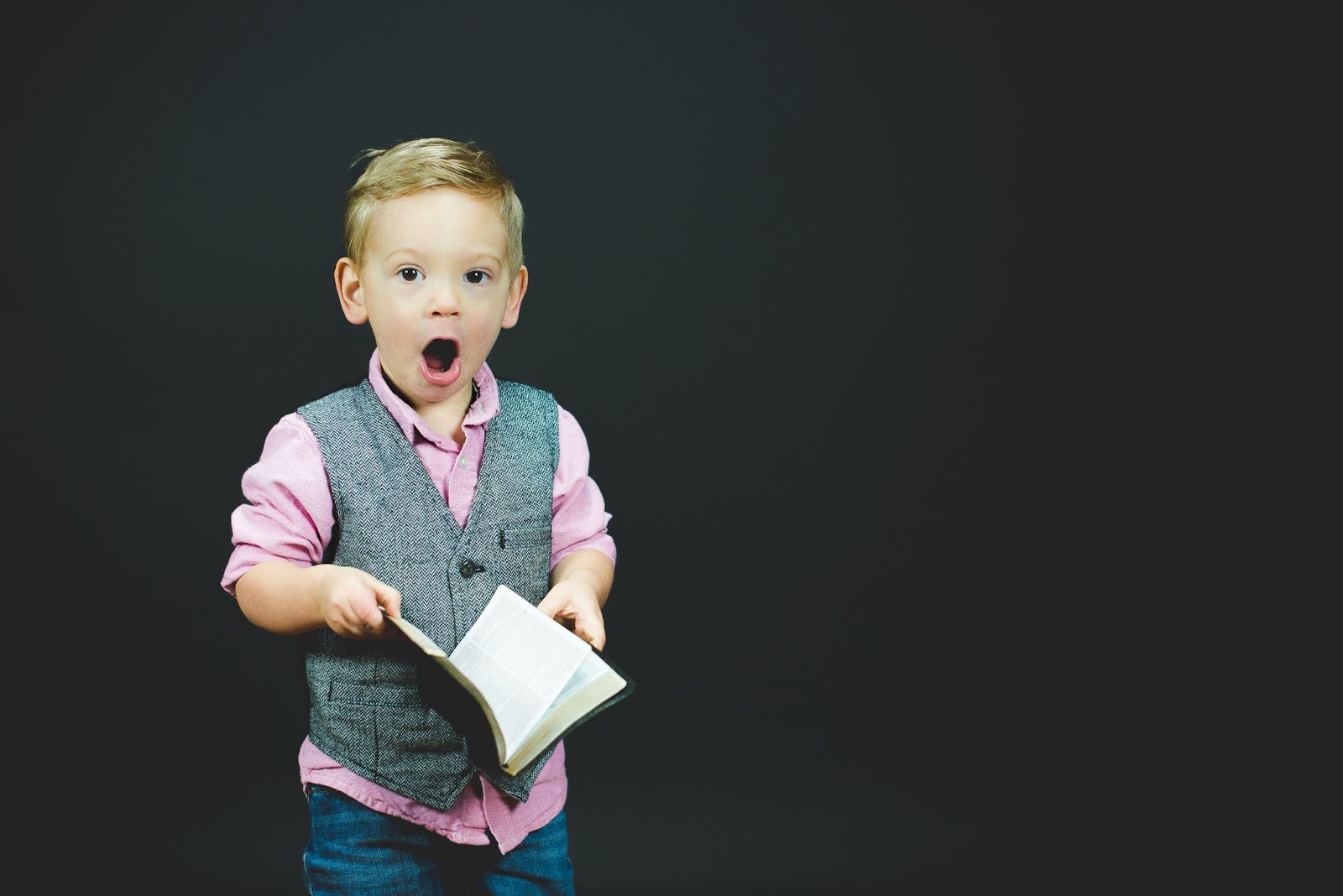 A young boy in a vest and pink shirt holds an open Bible with an expressive look of surprise and wonder, representing the joy of discovering God’s Word in children’s ministry.