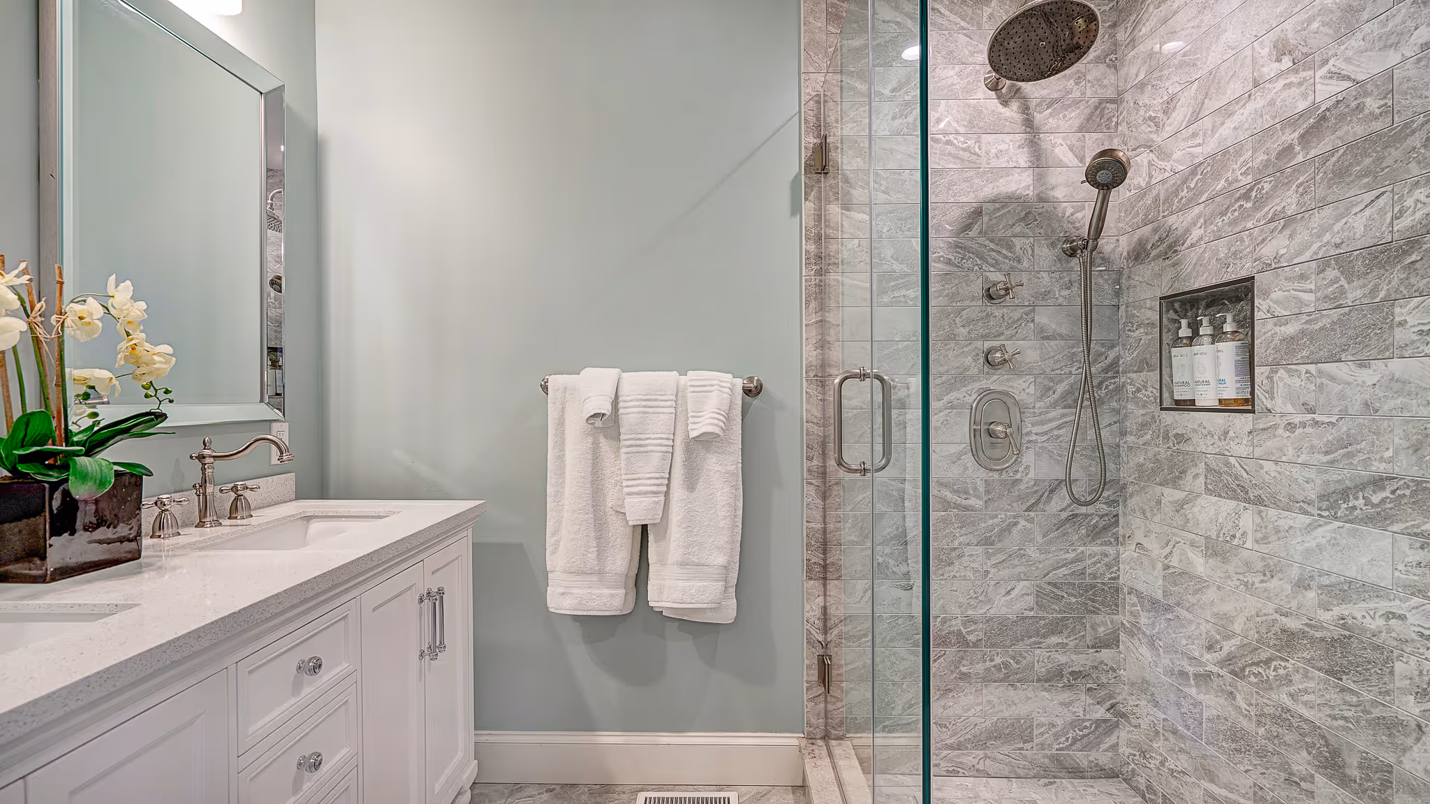 Modern bathroom with white vanity and countertop, mirror, white towels on a silver towel rack, and a glass-enclosed shower with gray marble tiles and mounted toiletries.