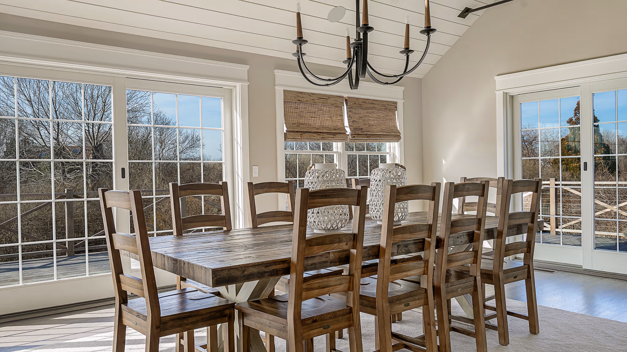 Rustic wooden dining table with eight matching chairs in a bright room with large windows and sliding glass doors.