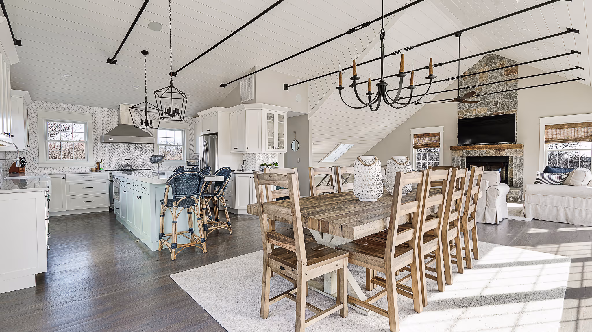 Open-concept dining and kitchen area with wooden dining table, chairs, island with blue bar stools, white cabinetry, and stone fireplace.