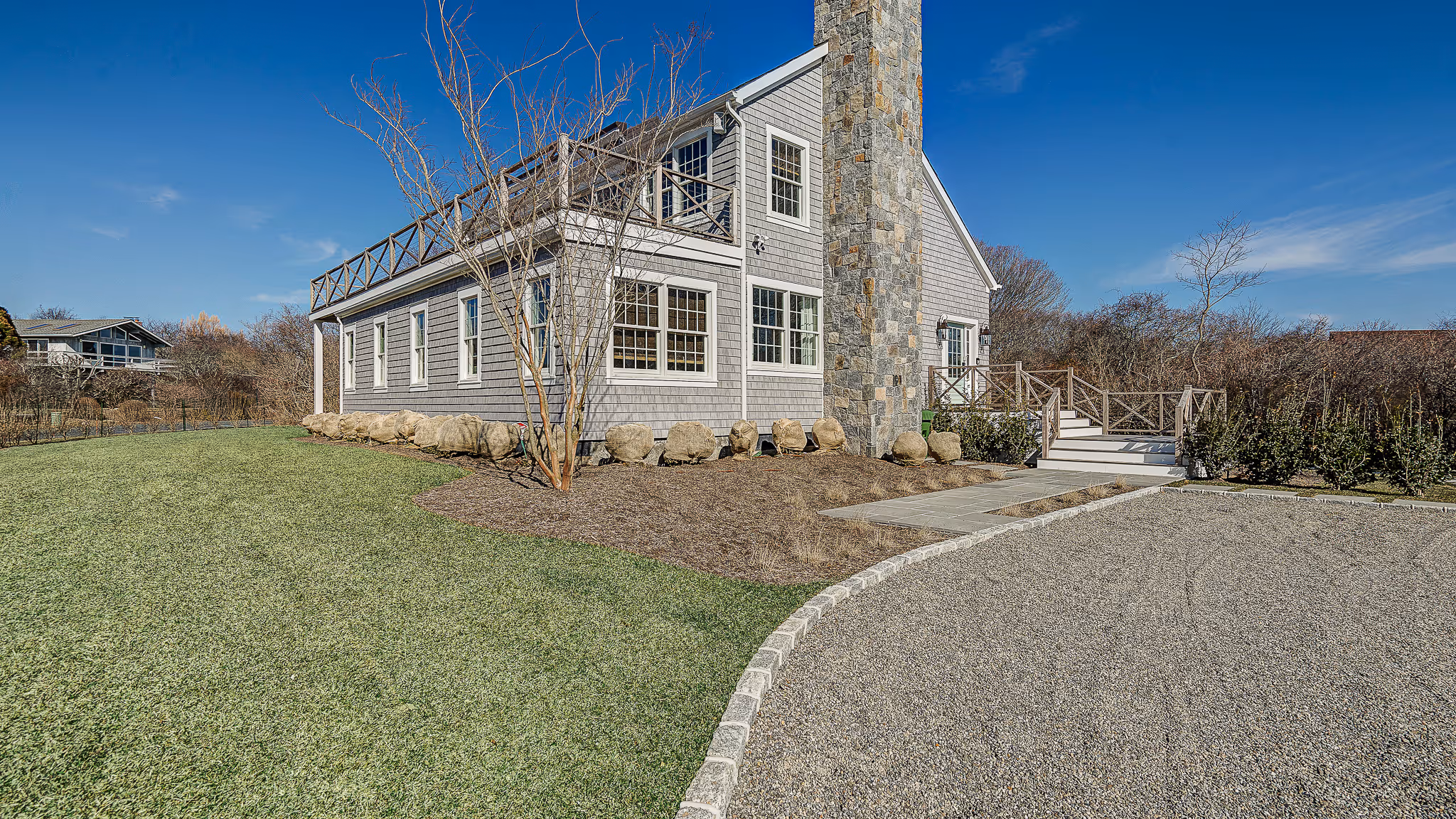 Gray two-story house with stone chimney, surrounded by a gravel driveway, green lawn, and leafless trees under a clear blue sky.