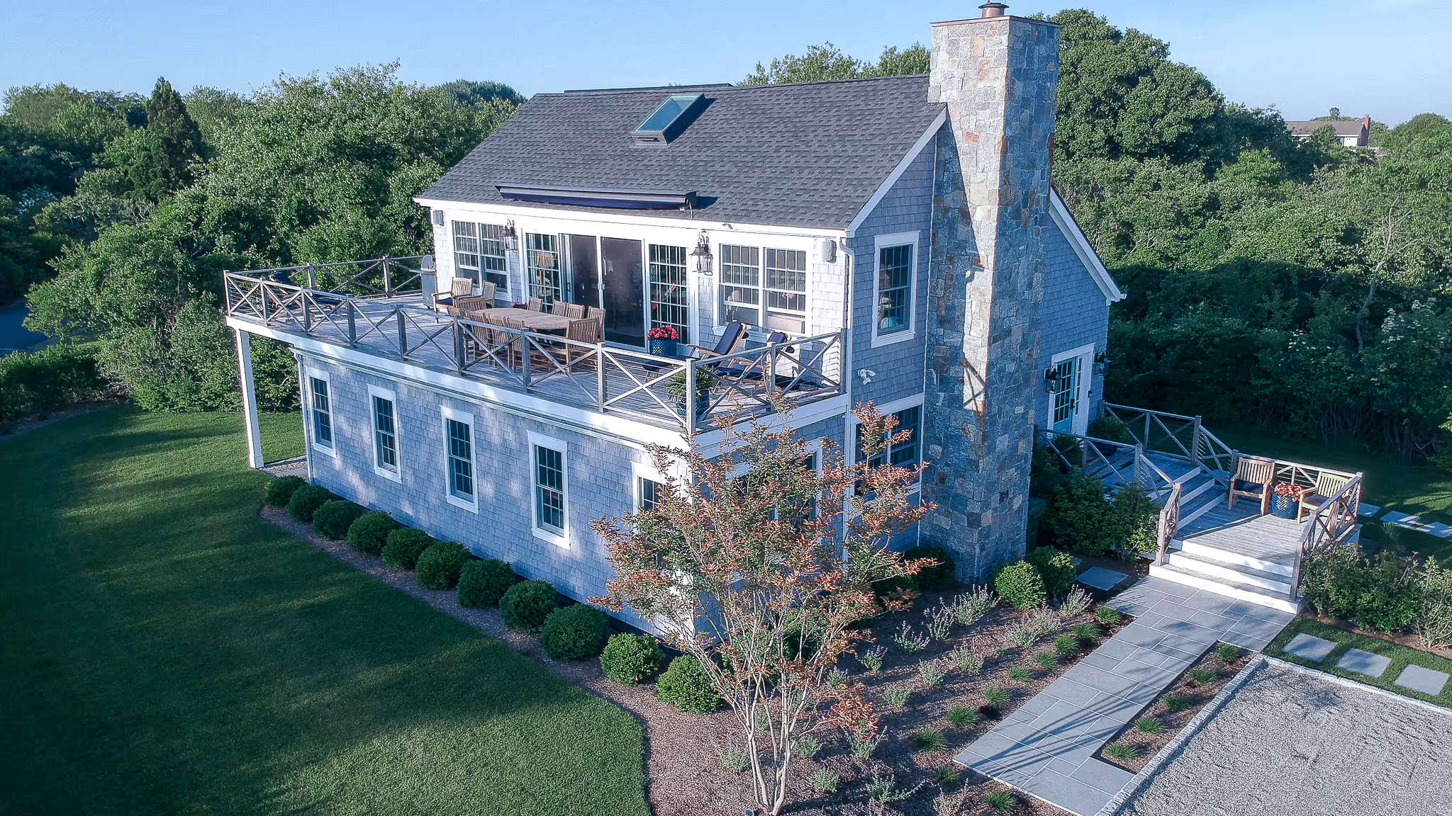 Two-story house with a large balcony featuring outdoor furniture and a stone chimney nestled among green trees and lawn.