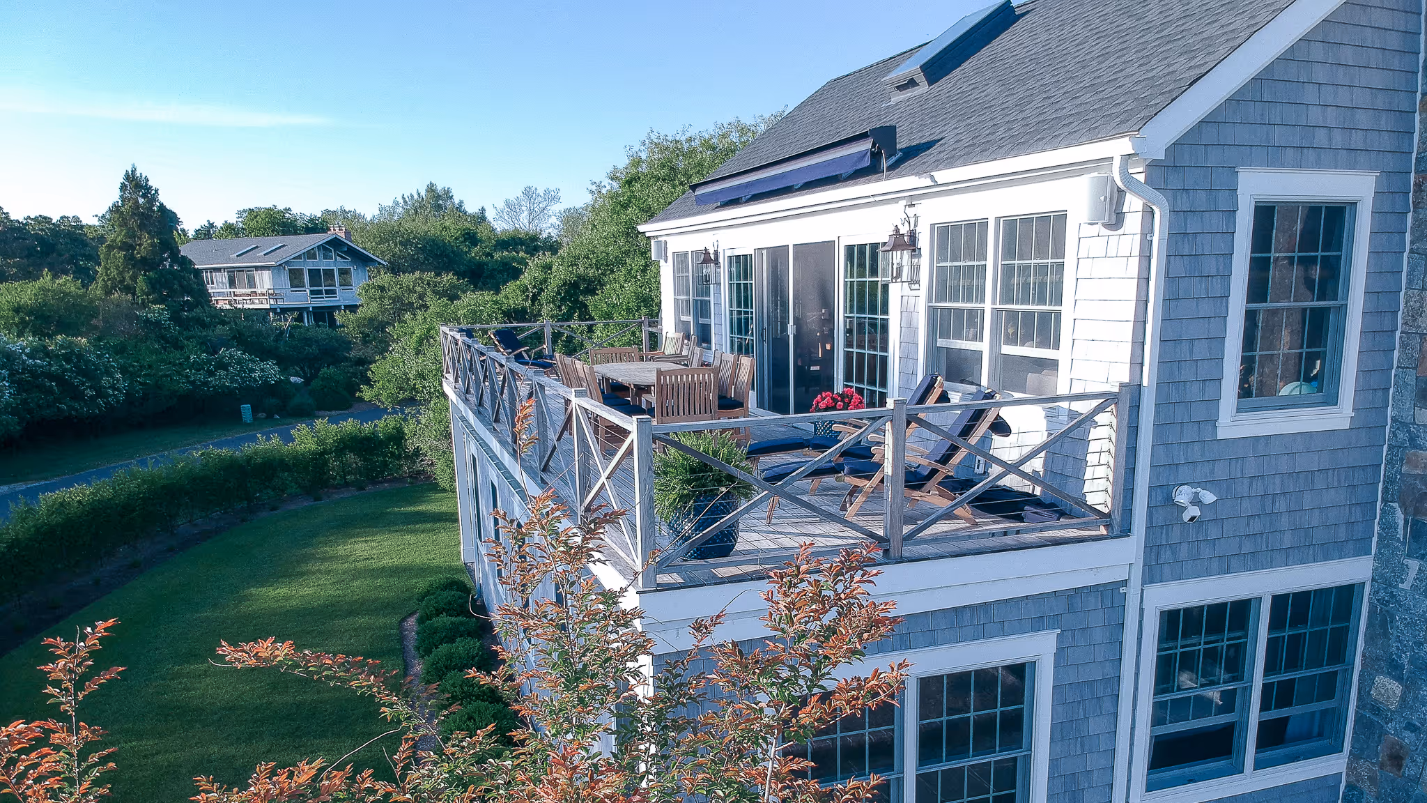 Elevated wooden deck with outdoor furniture attached to a gray two-story house surrounded by green trees and lawn.