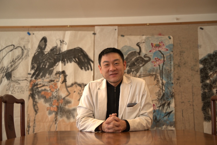 Xiao Yu Wang, L.Ac., seated at a table in front of traditional Chinese paintings